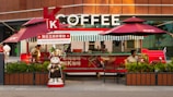 A coffee stand branded with KFC elements, including two large red umbrellas and a cartoon figure of a cat dressed in a barista outfit. The stand displays the word 'COFFEE' prominently in large letters. There are people working behind the counter, and potted plants are placed in front of the stand.