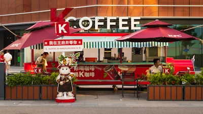 A coffee stand branded with KFC elements, including two large red umbrellas and a cartoon figure of a cat dressed in a barista outfit. The stand displays the word 'COFFEE' prominently in large letters. There are people working behind the counter, and potted plants are placed in front of the stand.