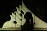 A modern abstract sculpture with curved and angular shapes, made of a light material, is set against a dark night background. The silhouette of a person appears in the foreground, adding a mysterious element to the scene.