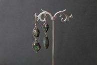 An elegant pair of earrings is displayed on a decorative silver stand. The earrings feature dark green gemstones set in intricate metal frames.