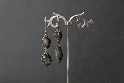 An elegant pair of earrings is displayed on a decorative silver stand. The earrings feature dark green gemstones set in intricate metal frames.