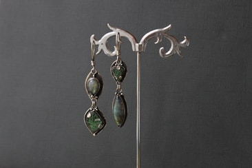An elegant pair of earrings is displayed on a decorative silver stand. The earrings feature dark green gemstones set in intricate metal frames.