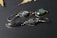 A pair of artisan earrings featuring polished stones set in silver. One earring displays a teardrop-shaped stone with iridescent hues, while the other has a triangular green stone with a textured surface. They are laid on a dark textured background with partial views of dried leaves in the corners, adding an organic touch.
