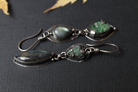 A pair of artisan earrings featuring polished stones set in silver. One earring displays a teardrop-shaped stone with iridescent hues, while the other has a triangular green stone with a textured surface. They are laid on a dark textured background with partial views of dried leaves in the corners, adding an organic touch.