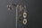 Two pairs of silver earrings with artistic and organic designs hang on a decorative metal stand against a dark textured background. The earrings feature hollow shapes and smooth, reflective surfaces.