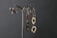 Two pairs of silver earrings with artistic and organic designs hang on a decorative metal stand against a dark textured background. The earrings feature hollow shapes and smooth, reflective surfaces.