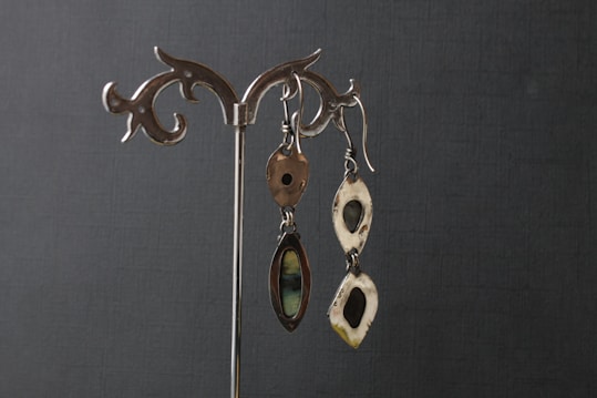 Two pairs of silver earrings with artistic and organic designs hang on a decorative metal stand against a dark textured background. The earrings feature hollow shapes and smooth, reflective surfaces.