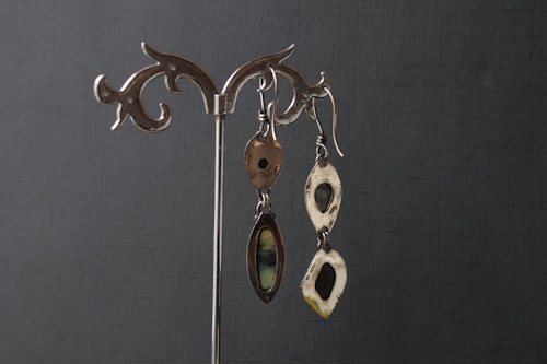 Two pairs of silver earrings with artistic and organic designs hang on a decorative metal stand against a dark textured background. The earrings feature hollow shapes and smooth, reflective surfaces.