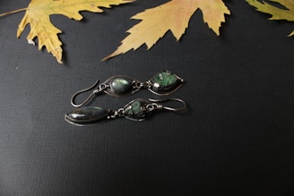 A pair of intricately designed silver earrings with dark green gemstones lies on a textured dark surface. Above them, dried yellow maple leaves are scattered, contrasting the dark background.