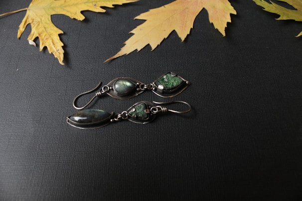 A pair of intricately designed silver earrings with dark green gemstones lies on a textured dark surface. Above them, dried yellow maple leaves are scattered, contrasting the dark background.