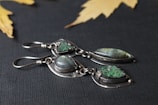 Two pairs of silver earrings set with green and iridescent stones are displayed on a dark textured background. The metal has a delicate, handcrafted look, and the stones are polished smooth, offering an elegant appearance. Yellow maple leaves are scattered in the blurred background, adding a natural touch to the composition.