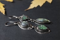 Two pairs of silver earrings set with green and iridescent stones are displayed on a dark textured background. The metal has a delicate, handcrafted look, and the stones are polished smooth, offering an elegant appearance. Yellow maple leaves are scattered in the blurred background, adding a natural touch to the composition.