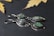 Two pairs of silver earrings set with green and iridescent stones are displayed on a dark textured background. The metal has a delicate, handcrafted look, and the stones are polished smooth, offering an elegant appearance. Yellow maple leaves are scattered in the blurred background, adding a natural touch to the composition.