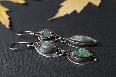 Two pairs of silver earrings set with green and iridescent stones are displayed on a dark textured background. The metal has a delicate, handcrafted look, and the stones are polished smooth, offering an elegant appearance. Yellow maple leaves are scattered in the blurred background, adding a natural touch to the composition.