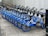 A row of blue and white rental bicycles are neatly parked in a designated area alongside a pathway. Each bike features a basket, handlebars, and a seat, aligned uniformly. The area is concrete-paved, with a metallic fence running parallel to the row of bicycles.