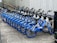 A row of blue and white rental bicycles are neatly parked in a designated area alongside a pathway. Each bike features a basket, handlebars, and a seat, aligned uniformly. The area is concrete-paved, with a metallic fence running parallel to the row of bicycles.