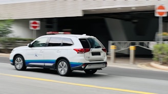 A white SUV with blue and red markings, possibly an emergency or official vehicle, is moving along a street. The background includes a building entrance, sidewalk, and some greenery. Traffic signs are visible, indicating a formal setting.