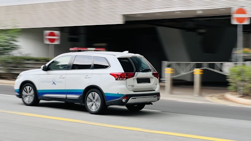A white SUV with blue and red markings, possibly an emergency or official vehicle, is moving along a street. The background includes a building entrance, sidewalk, and some greenery. Traffic signs are visible, indicating a formal setting.