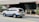 A white SUV with blue and red markings, possibly an emergency or official vehicle, is moving along a street. The background includes a building entrance, sidewalk, and some greenery. Traffic signs are visible, indicating a formal setting.