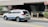 A white SUV with blue and red markings, possibly an emergency or official vehicle, is moving along a street. The background includes a building entrance, sidewalk, and some greenery. Traffic signs are visible, indicating a formal setting.