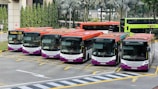 Several buses in different colors are parked in a bus depot. The buses are arranged side by side, with visible signs indicating the route number 232. Palm trees and a building are in the background.