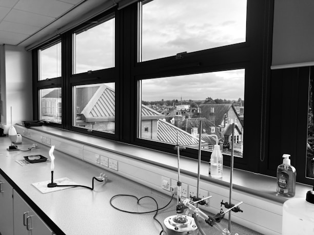 A laboratory setting with a long counter space equipped with various scientific instruments and equipment. There are multiple large windows offering a clear view of the rooftops and buildings outside. On the counter, there is a lit Bunsen burner, a lab water bottle, and a bottle of hand sanitizer among other lab apparatus.