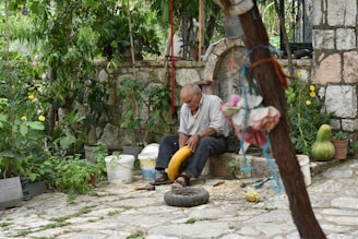 An elderly man sitting peacefully in a garden with robotic companions tending the plants around him.