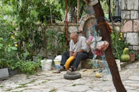 An elderly man is seated on a stone bench in a garden setting, surrounded by various plants and flowers. He appears to be focused on a large yellow squash or gourd in his hands. Nearby, there are several plastic buckets, gardening tools, and a few scattered items on the ground. The setting includes a rustic stone wall and path, giving a serene and old-fashioned atmosphere.