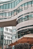 A modern building with curved glass and stone elements, featuring a unique architectural design. The structure has large windows and is connected by an overhanging bridge section. In the foreground, there is an outdoor seating area with a beige canopy.