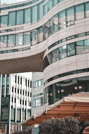A modern building with curved glass and stone elements, featuring a unique architectural design. The structure has large windows and is connected by an overhanging bridge section. In the foreground, there is an outdoor seating area with a beige canopy.