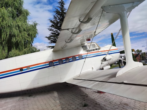 A large, vintage-style biplane is positioned on a paved area. The aircraft is primarily white, with decorative blue and orange horizontal stripes along its body. Lush green trees and a partly cloudy blue sky form the backdrop, creating a picturesque outdoor setting.