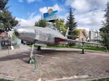 A vintage aircraft is displayed outdoors, resting on a paved area surrounded by a low barrier. The aircraft has a sleek, metallic body with the number 8733 marked on the side. Trees and buildings are visible in the background, along with a sign written in a different language.
