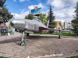 A vintage aircraft is displayed outdoors, resting on a paved area surrounded by a low barrier. The aircraft has a sleek, metallic body with the number 8733 marked on the side. Trees and buildings are visible in the background, along with a sign written in a different language.
