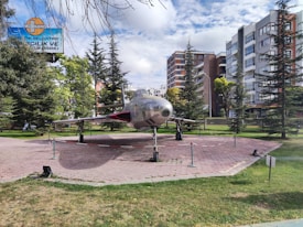 An aircraft displayed outdoors in a park surrounded by trees and adjacent to multi-story buildings. The aircraft is mounted on a paved area with a barrier around it, and a sign in the background suggests the park's name.