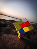 A partially scrambled Rubik's Cube is placed on a rock by the shore. The background features a calm body of water under a dusky sky, transitioning from light to dark as it meets the horizon.