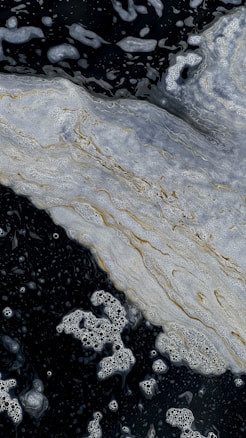 Swirling patterns of foam and bubbles float on a dark liquid surface, creating an abstract and dynamic scene. The foam appears to have intricate textures and layers, with hints of gold and white blending seamlessly.