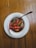 A small white bowl containing different kinds of chili peppers, including red and green ones, soaked in a dark liquid. The bowl is placed on a white saucer, and a metal spoon rests inside the bowl. The setup is placed on a wooden table.
