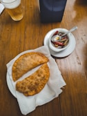 Two fried empanadas sit on a white plate lined with a paper napkin on a wooden table. Next to them, a small bowl contains pickled peppers and a spoon. A glass of beer and an opaque blue object are partially visible near the top of the image.