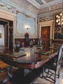 An ornate and historical meeting room features a large, polished wooden table surrounded by high-backed chairs. Detailed woodwork and sculptures adorn the walls and ceiling. A large, framed painting depicting several individuals in formal attire is prominently displayed. The room is lit by an elegant chandelier and wall sconces, highlighting the room's classic and sophisticated decor.
