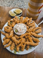 A plate of golden, crispy fried food with two forks is centered on a wooden table. In the middle of the plate, a bowl of creamy sauce filled with vegetables sits atop shredded greens. Sliced limes on a separate dish are positioned behind the plate. A striped yellow and red container is also visible in the background.