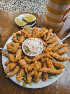 A plate of golden, crispy fried food with two forks is centered on a wooden table. In the middle of the plate, a bowl of creamy sauce filled with vegetables sits atop shredded greens. Sliced limes on a separate dish are positioned behind the plate. A striped yellow and red container is also visible in the background.
