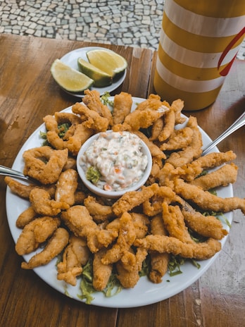 A plate of golden, crispy fried food with two forks is centered on a wooden table. In the middle of the plate, a bowl of creamy sauce filled with vegetables sits atop shredded greens. Sliced limes on a separate dish are positioned behind the plate. A striped yellow and red container is also visible in the background.