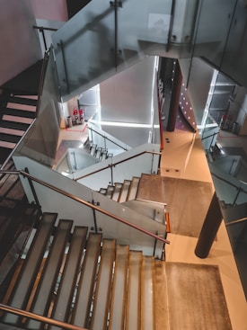 A modern indoor staircase with glass railings and a sleek, architectural design. The stairs spiral down, creating geometric patterns. Fire extinguishers are visible at one of the landings, and there are bright accent lights illuminating the space.