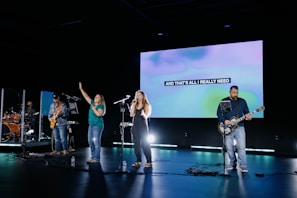 A band performs on stage with five members, including a drummer, guitarists, and vocalists. The stage is lit with spotlights, and a large screen in the background displays the text 'AND THAT'S ALL I REALLY NEED'. The atmosphere suggests a live music performance or rehearsal.