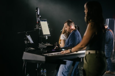 A group of musicians is performing on stage. The focus is on a keyboard player in the foreground, with another musician visible in the background playing a different instrument. The lighting is dim and atmospheric, creating an intimate concert setting. There are several electronic devices and musical equipment visible around the musicians.