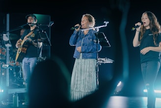 A musical performance with three individuals on stage, one playing a guitar and two others singing into microphones. Stage lighting casts a dynamic atmosphere, highlighting the performers. A silhouette of an audience member raises their hand in the foreground.