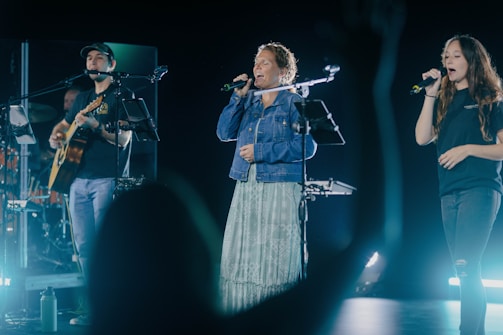 A musical performance with three individuals on stage, one playing a guitar and two others singing into microphones. Stage lighting casts a dynamic atmosphere, highlighting the performers. A silhouette of an audience member raises their hand in the foreground.