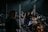 A live music performance featuring several musicians on stage. One person is enthusiastically singing into a microphone, while another plays the guitar. The setup includes a drum set and various microphones, suggesting a band setting. The lighting is dim with spotlights focused on the performers, creating a concert-like atmosphere.