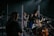 A live music performance featuring several musicians on stage. One person is enthusiastically singing into a microphone, while another plays the guitar. The setup includes a drum set and various microphones, suggesting a band setting. The lighting is dim with spotlights focused on the performers, creating a concert-like atmosphere.