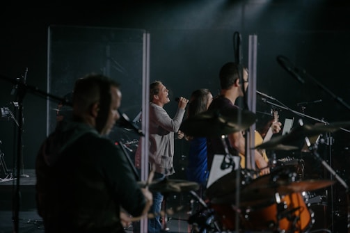 A live music performance featuring several musicians on stage. One person is enthusiastically singing into a microphone, while another plays the guitar. The setup includes a drum set and various microphones, suggesting a band setting. The lighting is dim with spotlights focused on the performers, creating a concert-like atmosphere.